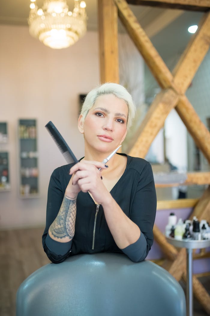 Blonde female hairstylist with tattoos holding comb and scissors in a chic salon setting.